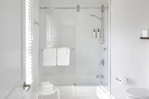 A modern bathroom with a glass-enclosed shower, white tiles, a toilet, and a small white stool.