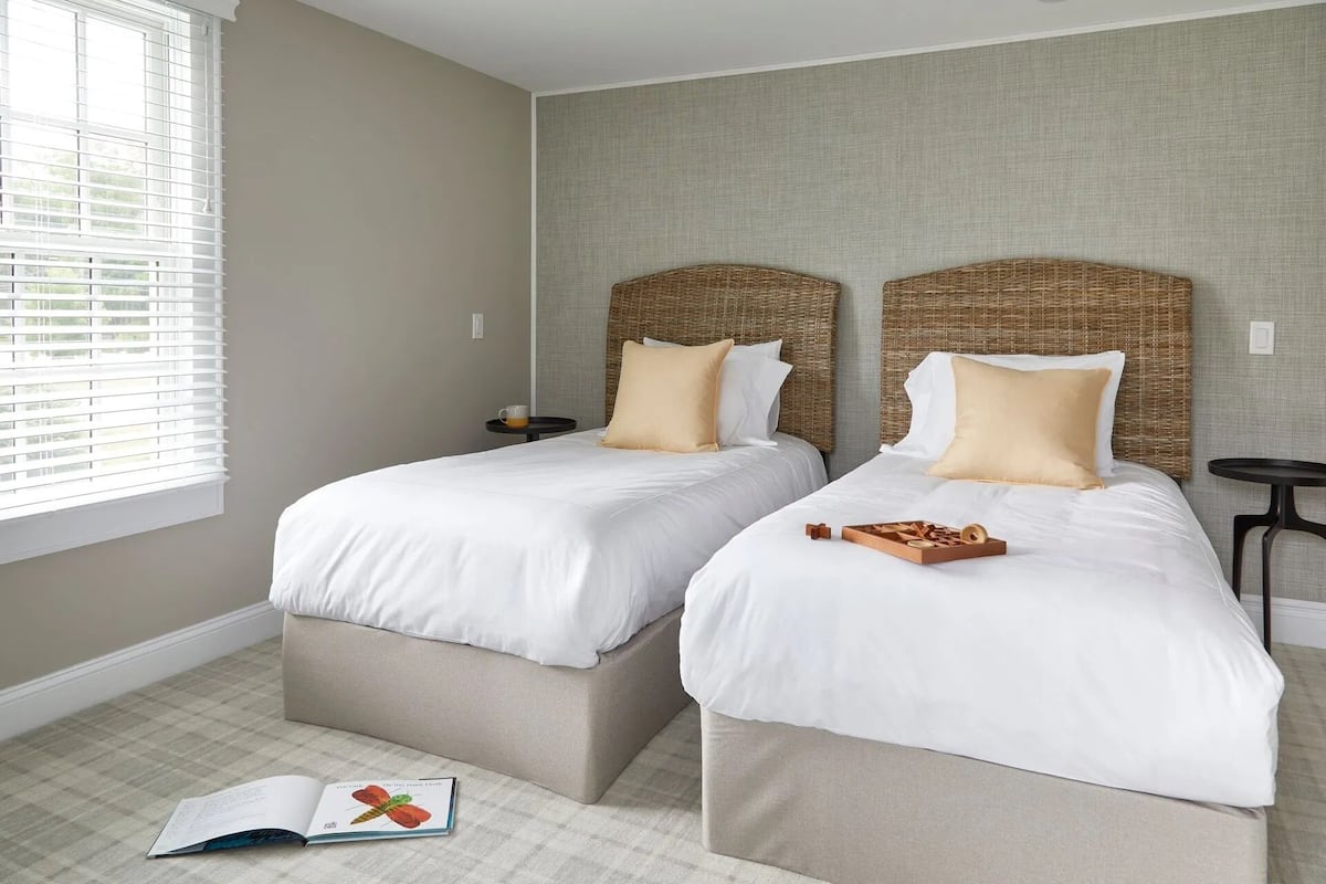 A hotel room with two beds, a small table, and a window with blinds.