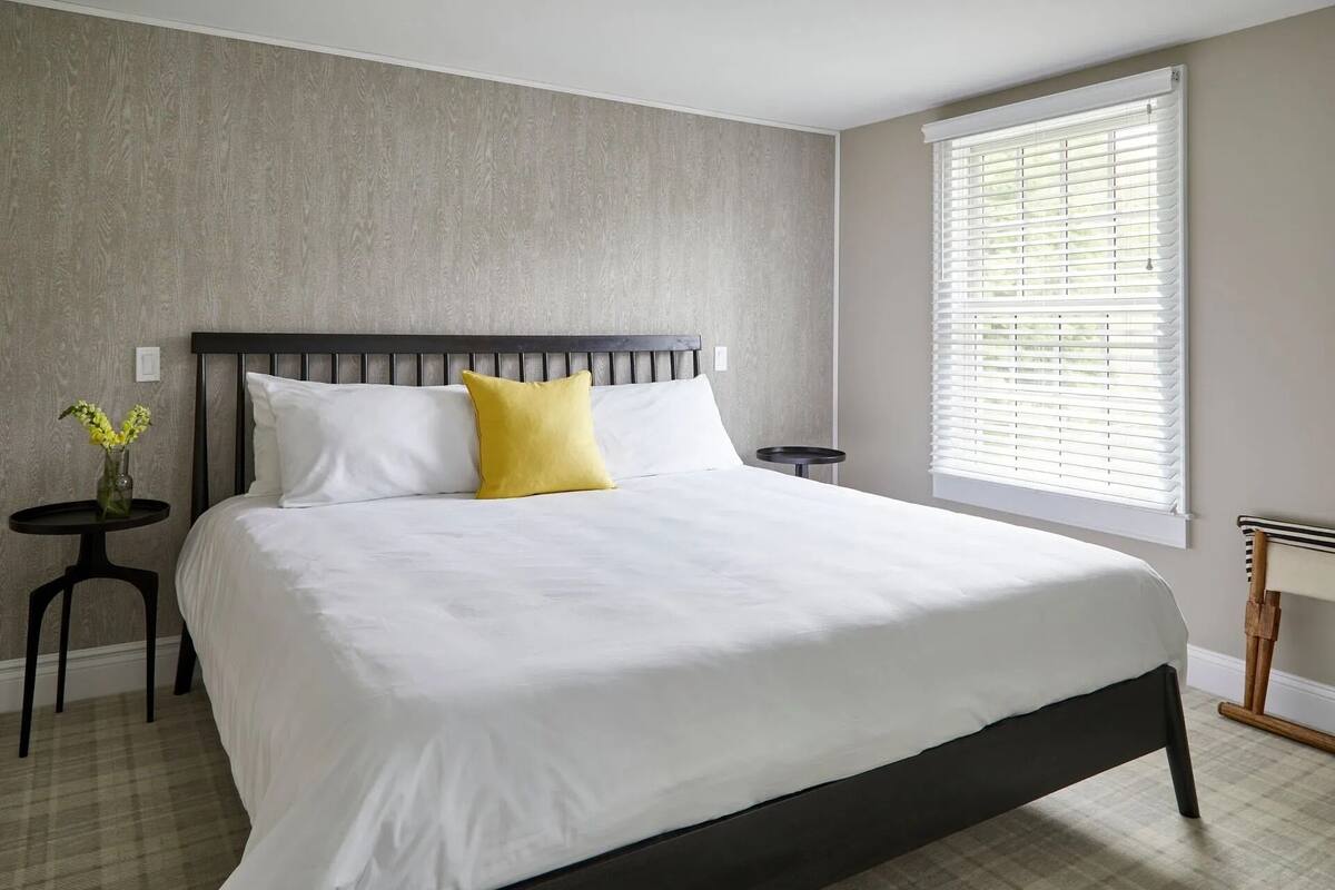 A bedroom with a large bed, a nightstand, a small table with a vase, and a window with blinds.
