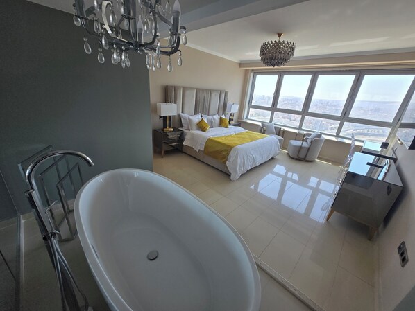 A modern bedroom with a large bathtub, a bed with a yellow accent, and a view of the city through large windows.