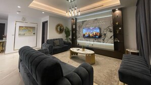 A modern living room with a large flat-screen TV, dark sofas, a marble accent wall, and a ceiling light fixture. - 2beroom Apartments in Eko Atlantic City (Lagos)