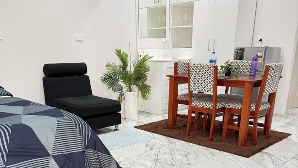 A modern living room with a bed, a black sofa, a dining table with chairs, a potted plant, and a kitchen area with cabinets and a refrigerator. - Cosy self contained studio apartment,5 mins from Nadi International Airport. (Nadi)