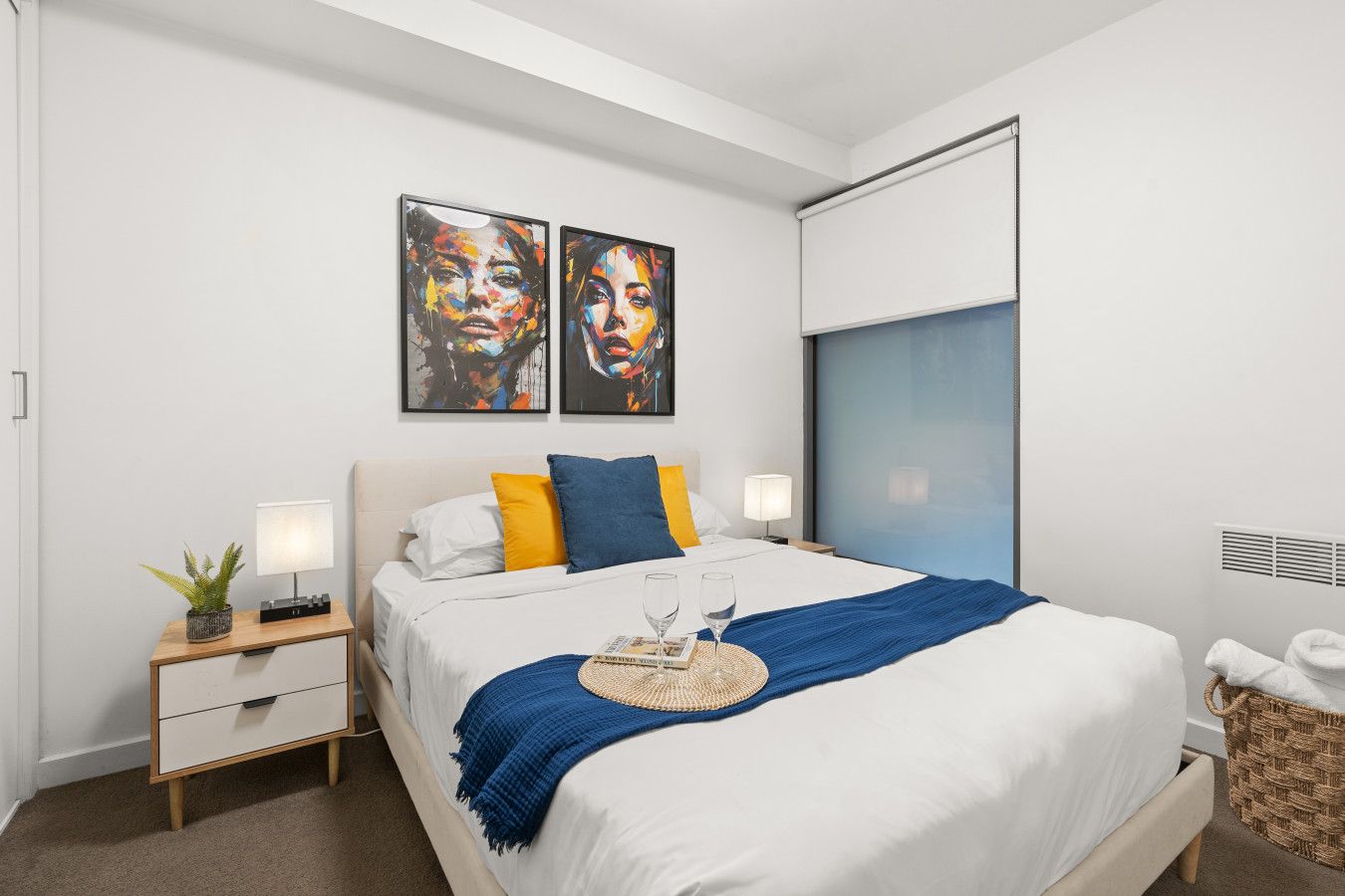Una habitación de hotel moderna con una cama grande, dos mesitas de noche, una planta pequeña en maceta y dos cuadros abstractos en la pared.