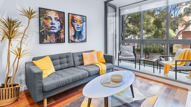 A modern living room with a grey sofa, yellow cushions, a round table, and large windows offering a view of the outdoors.