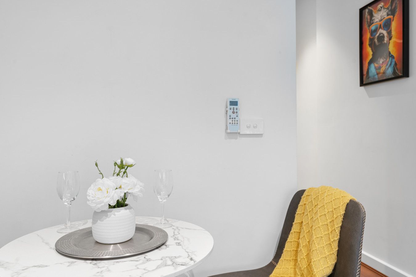 Una habitación moderna con una mesa de mármol, un jarrón blanco con flores, dos copas de vino, una silla con una manta amarilla y un cuadro en la pared.