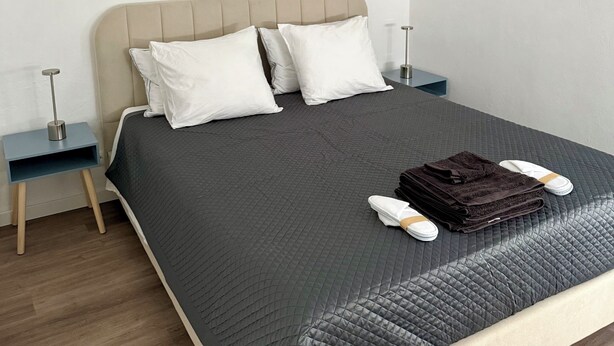 Een keurig opgemaakt bed met een grijze deken, twee witte kussens en een bruine handdoek gevouwen op het voeteneinde. Aan beide zijden van het bed staan nachtkastjes met lampen.