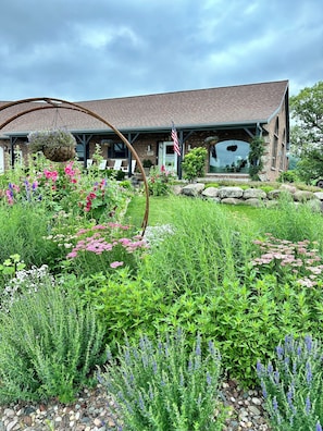 Ein rustikales Backsteinhaus mit steinernen Fundamenten, umgeben von einem gepflegten Garten mit verschiedenen blühenden Pflanzen und einem hängenden Blumenkorb.