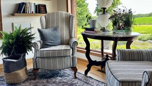 Une pièce chaleureuse avec un fauteuil à rayures, un pouf assorti, un tapis à motifs, une bibliothèque en bois agrémentée d’objets décoratifs et une grande fenêtre donnant sur un paysage verdoyant.