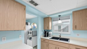 A modern kitchen with light blue walls, wooden cabinets, a built-in microwave, a coffee maker, and a window with a view of a staircase. - Walk To Beach, North End Restaurants, Night Life Steps Away! (Fort Myers Beach)