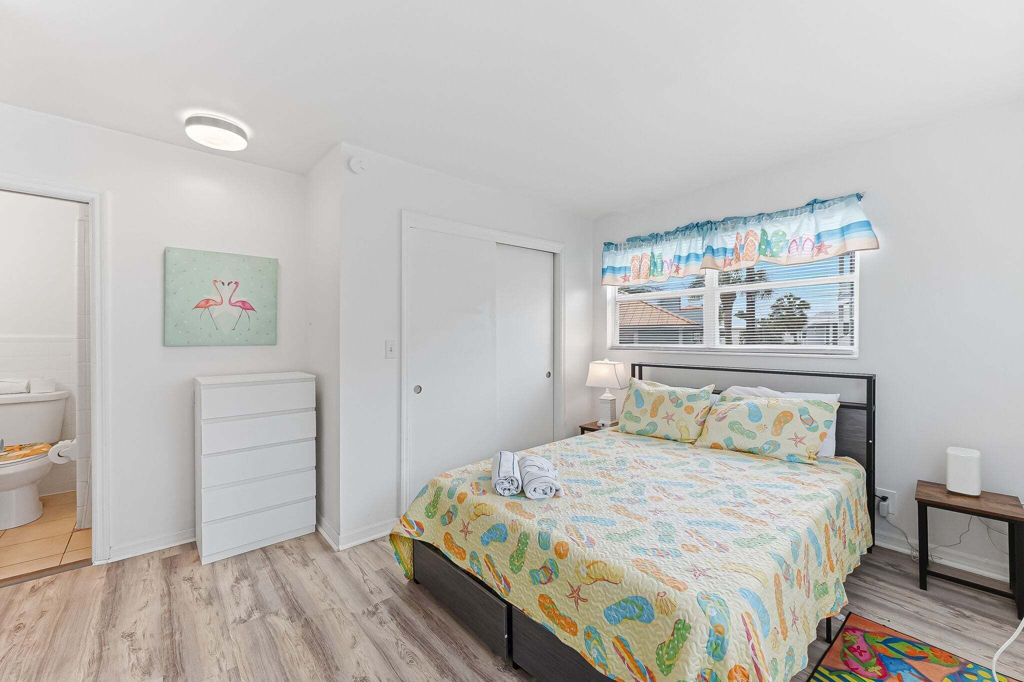 A bedroom with a bed, a bedside table, a nightstand, a window with a curtain, a wall-mounted air conditioning unit, and a small rug.