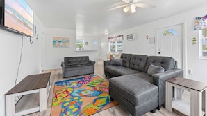 A living room with a gray sectional sofa, a small table, a colorful rug, a ceiling fan, and a TV on the wall.