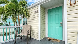 A turquoise door with a white frame, a number '2621' sign, a wicker chair, and a palm tree on a wooden porch. - Walk To Beach, North End Restaurants, Night Life Steps Away! (Fort Myers Beach)