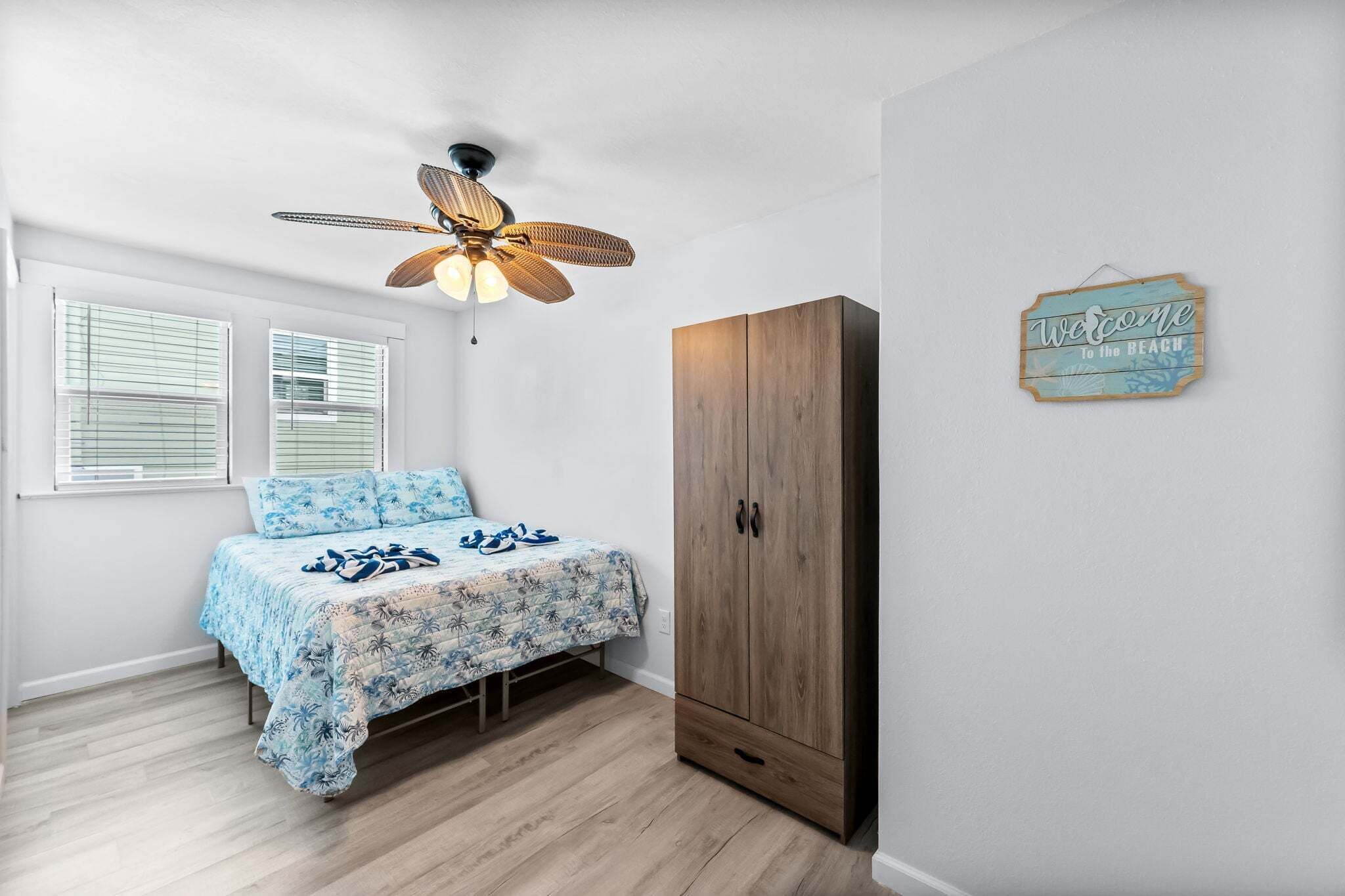 Une chambre à coucher avec un lit, une armoire en bois, un ventilateur de plafond et une pancarte indiquant « Bienvenue à la plage ».