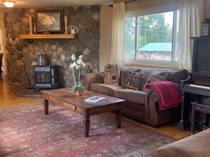 A cozy living room with a stone wall, a fireplace, a sofa with patterned cushions, a coffee table with a book, and a piano.