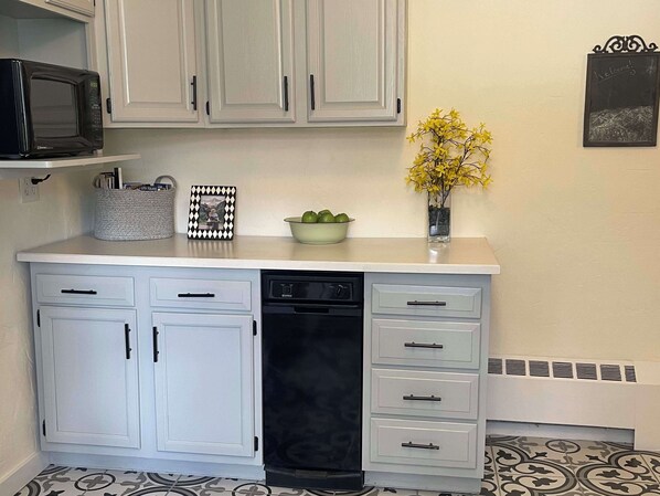 A kitchen with white cabinets, a dishwasher, a microwave, a bowl of green apples, and a vase with yellow flowers.