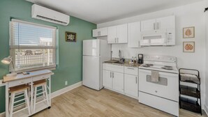 A compact kitchen with white appliances, a window with blinds, and a small desk with a chair.