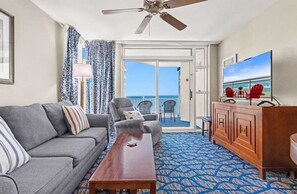 Une pièce comprenant un canapé, une table basse, un téléviseur, un ventilateur de plafond et une vue sur l’océan, accessible par une porte coulissante.