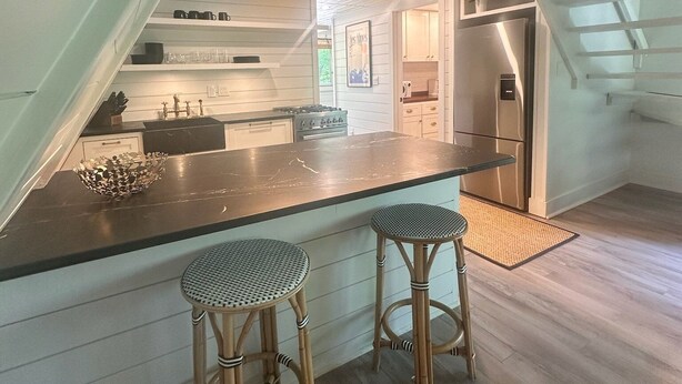 A kitchen with a central island, two bar stools, pendant lights, and a view of the living area through an open archway.