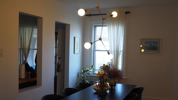 A dining area with a long table, chairs, a vase with flowers, and a ceiling light fixture.