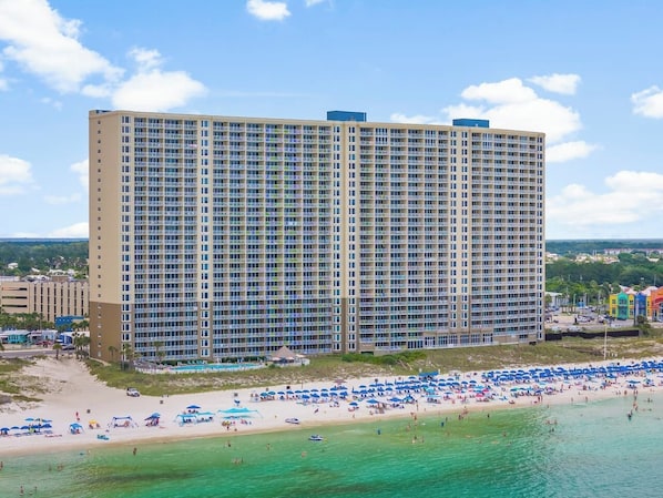 Un hotel grande con varios pisos y balcones, una playa con numerosas sombrillas y una vista despejada del océano.