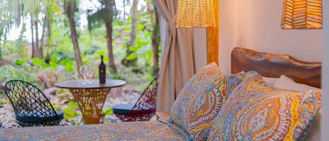 A bed with colorful patterned bedding, a woven light fixture, and a view of lush greenery outside.