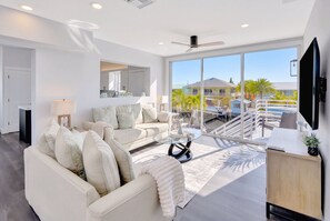 A modern living room with a sofa, coffee table, and a large window offering a view of a pool and buildings.