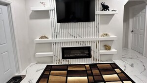 A modern living room with a flat-screen TV mounted on a white, vertically paneled wall. There are white shelves with decorative items and a patterned rug on the floor.