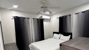 A bedroom with a bed, a ceiling fan, an air conditioning unit, and a wooden nightstand.