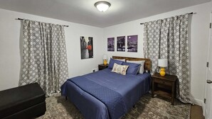 Une chambre avec un grand lit, deux tables de chevet avec des lampes, des rideaux à motifs et une œuvre d’art représentant un paysage urbain accrochée au mur.