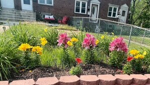 Un immeuble résidentiel à la façade en briques, doté de nombreuses fenêtres et d’un jardin devant avec des fleurs colorées et une clôture.