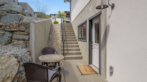 Un estrecho pasillo exterior con un muro de piedra, una pequeña mesa redonda y dos sillas de mimbre, y una escalera que lleva a una puerta con ventana.