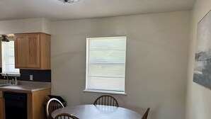A dining area with a round table and four chairs, a kitchen with wooden cabinets, and a window with blinds.
