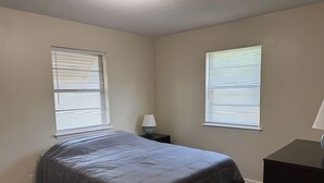 A bedroom with a bed, two windows with blinds, a nightstand with a lamp, and a dresser.