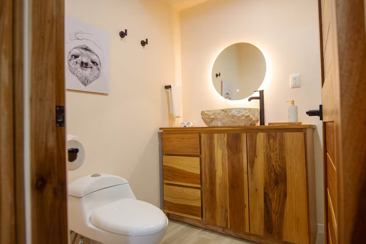 Un baño con inodoro blanco, tocador de madera y un espejo redondo.