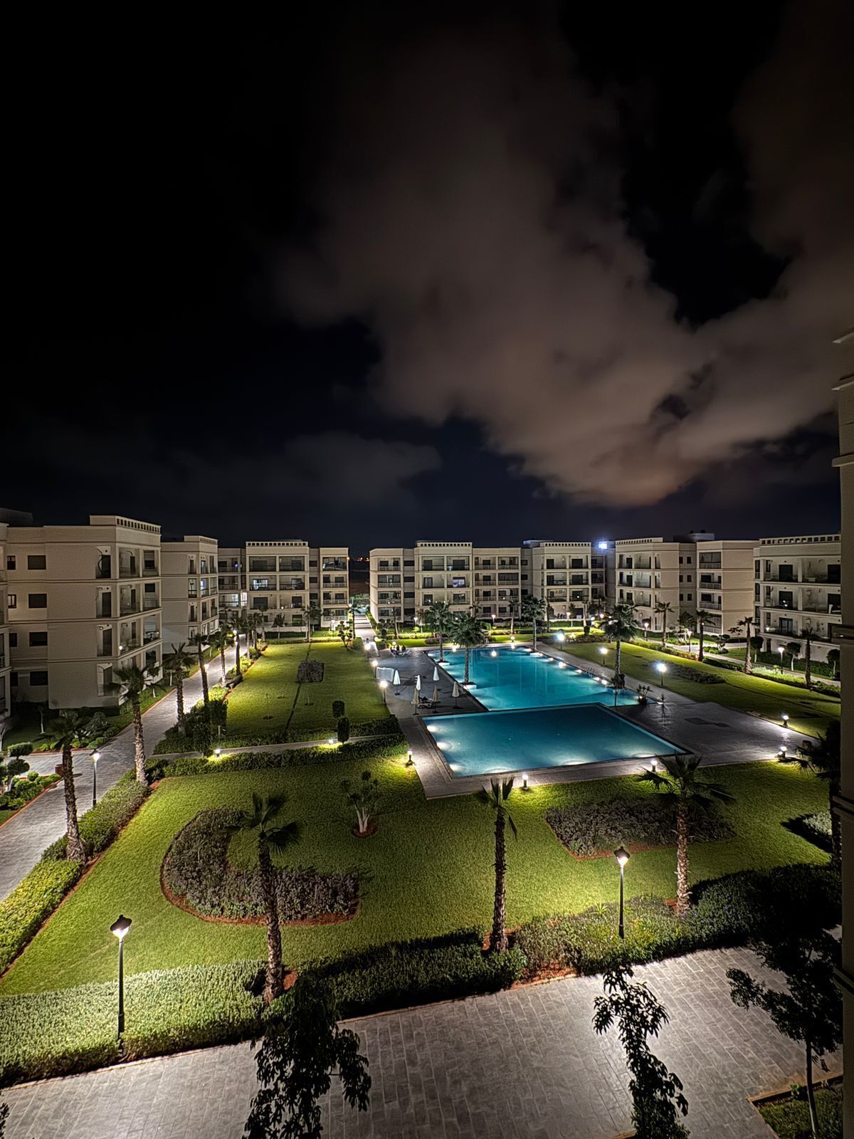 Ein gut beleuchteter Pool, umgeben von ordentlich gestutztem Rasen und Palmen, mit modernen Gebäuden im Hintergrund.