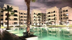 Un hôtel à plusieurs étages doté d’une piscine, de palmiers et de balcons bien éclairés.
