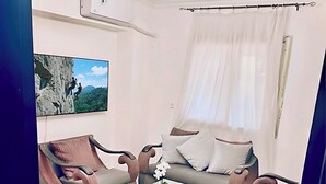 Un salón moderno con televisor de pantalla plana, ventilador de techo y un ventanal con cortinas translúcidas.