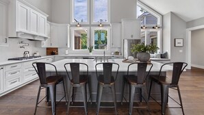 Una cocina amplia con una isla grande, armarios blancos y vistas al exterior a través de ventanas amplias.
