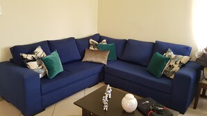 A blue sectional sofa with patterned cushions, a small black table with figurines and a vase, and a white lamp. - Warm house, away from the noise of the city. (Santa Cruz de la Sierra)