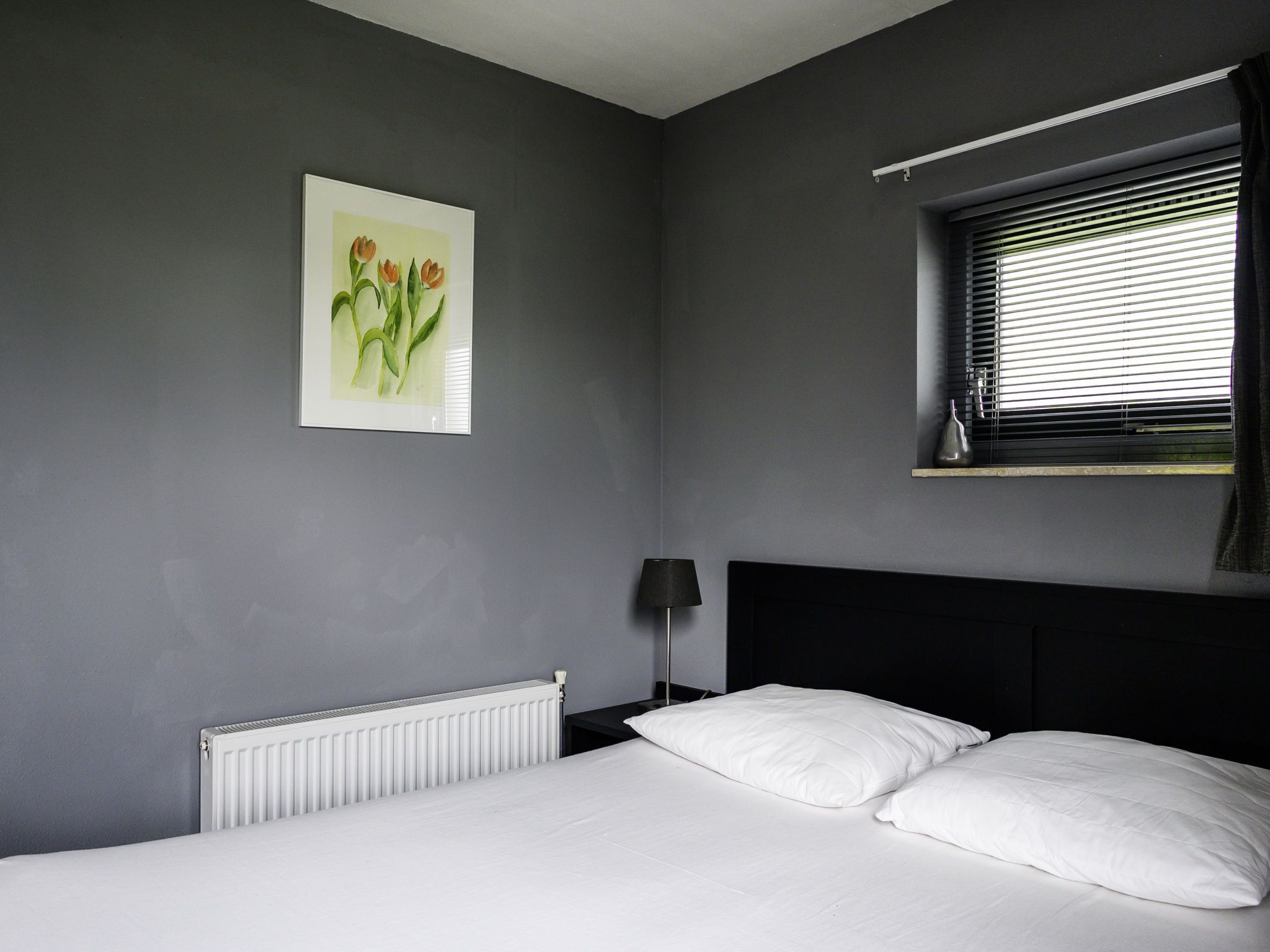 A bedroom with a bed, a radiator, a nightstand, a lamp, a window with blinds, and a painting on the wall.