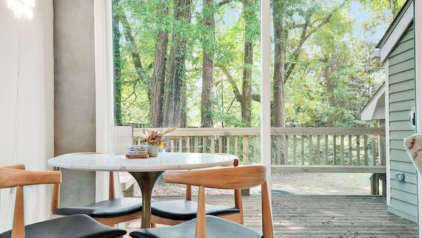 Un comedor con una mesa redonda y cuatro sillas de madera con cojines negros, una ventana con vista a una terraza de madera y árboles, y un pequeño florero con flores sobre la mesa.