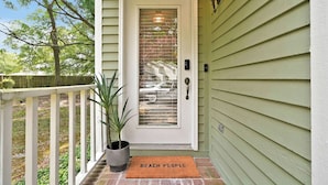 Una casa con fachada verde, una puerta blanca con un panel de vidrio, una planta en maceta y un tapete con la inscripción 'BEACH PEOPLE'.