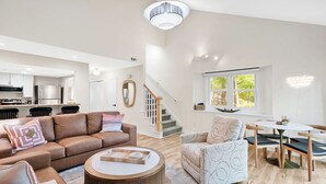 Amplia sala de estar con un sofá de cuero marrón, una mesa de centro redonda de madera y un sillón estampado. Una escalera con barandilla de madera conduce a una zona de cocina al fondo.