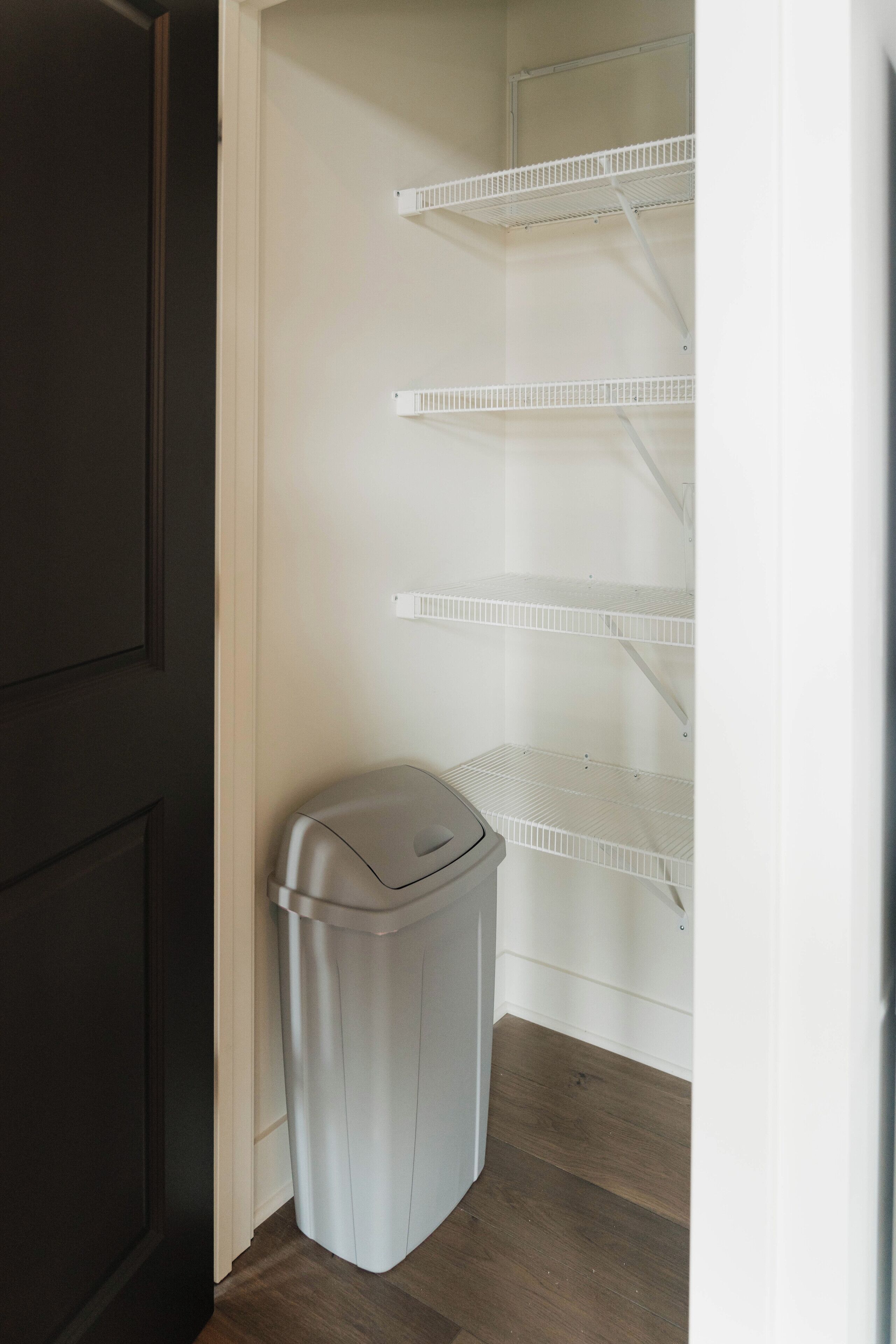 A laundry room with a trash bin and white shelving units.