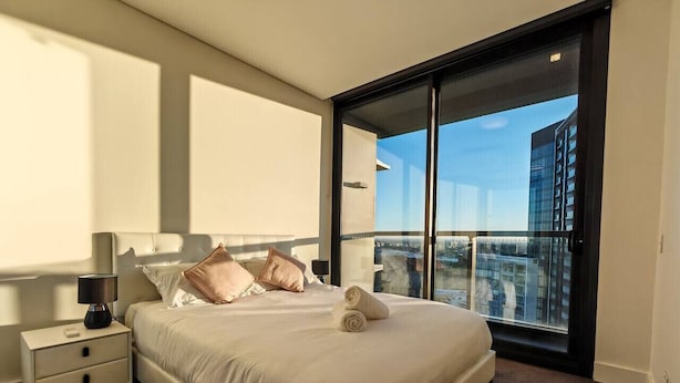 Un dormitorio moderno con una cama grande, una mesita de noche y vistas a un paisaje urbano a través de un ventanal corredero.