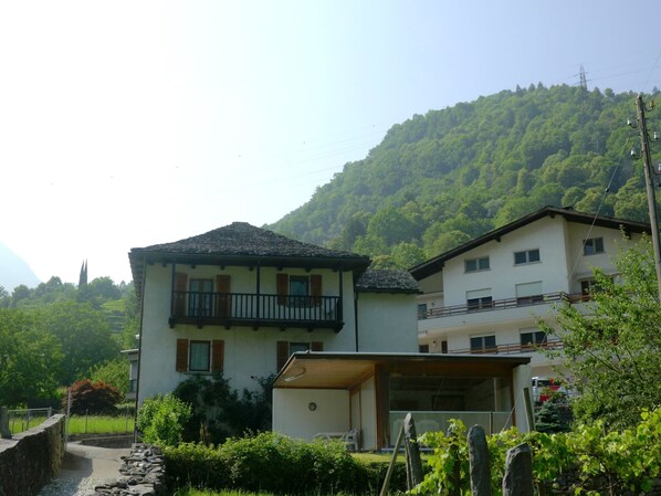 Um edifício de dois andares com varanda, cercado por vegetação e com montanhas ao fundo.