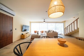 Een moderne woonkamer met een houten eettafel, een grote geweven lamp, een zithoek en een trap.