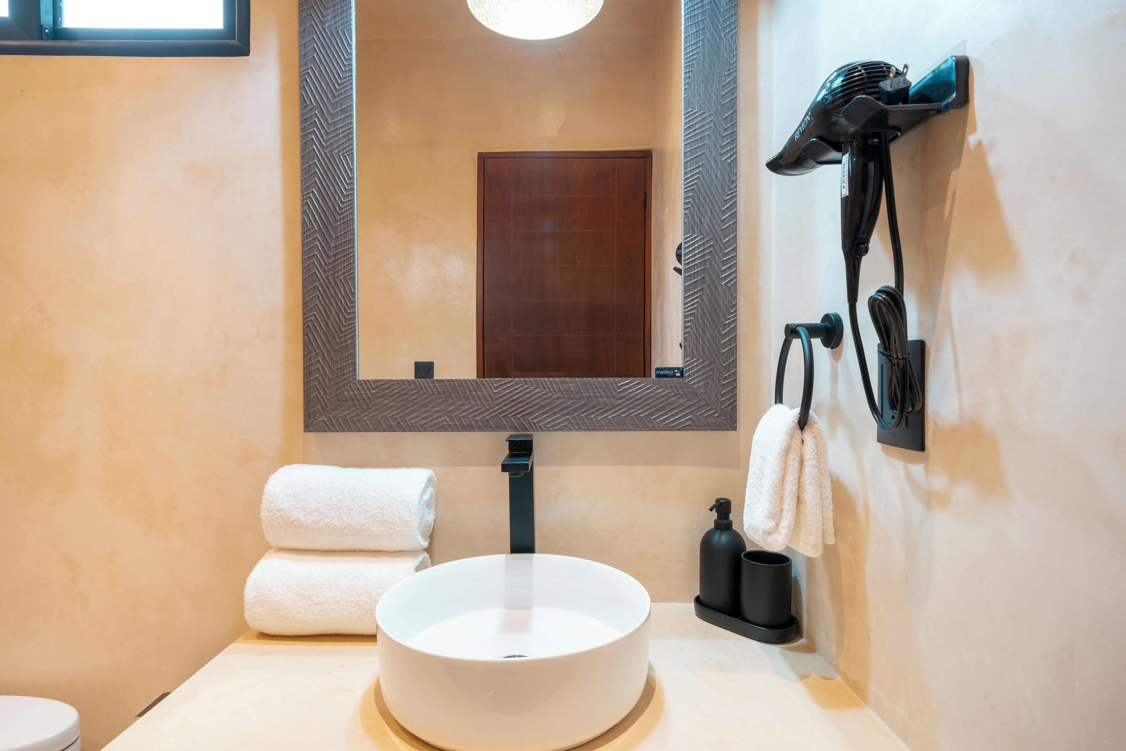 Un baño con un lavabo redondo, un espejo, un perchero para toallas y un secador de pelo montado en la pared.