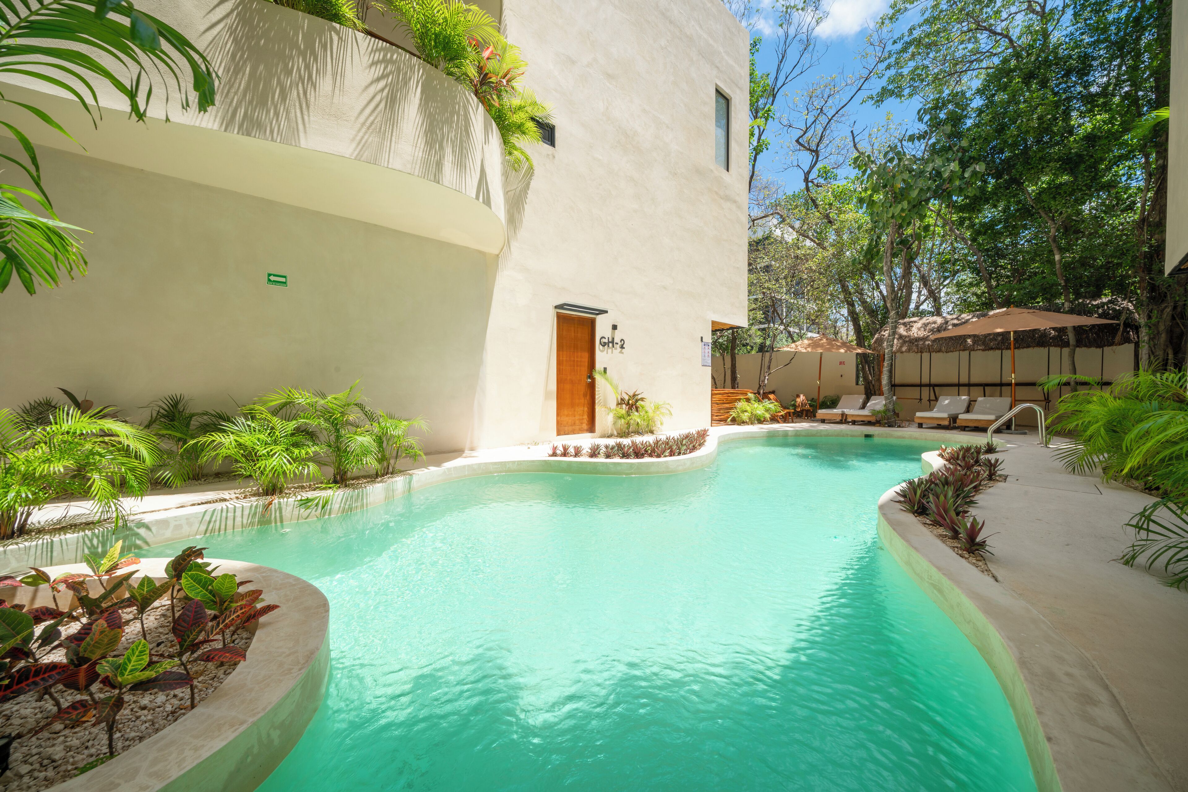 Un espace piscine à bords incurvés, entouré de plantes et bordé d’un bâtiment dont la porte est marquée du numéro « GH-2 ».