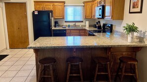 Una cocina con armarios de madera, encimera de granito, refrigerador, estufa y lavaplatos. Hay tres taburetes de madera y una planta en maceta.